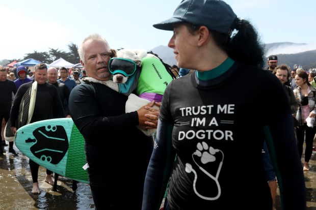 James Wall gets an escort while carrying Faith down to the shore to compete in the World Dog Surfing Championships, Saturday, Aug. 5, 2023, at Linda Mar Beach in Pacifica, Calif. (Karl Mondon/Bay Area News Group)