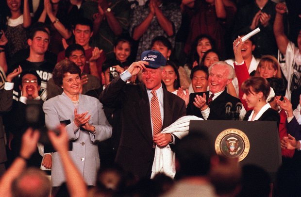 Then-President Bill Clinton dons a Carlmont cap from student Therese Sangervasi at a lectern with Sen. Dianne Feinstein and Rep. Tom Lantos at Carlmont High School in Belmont, Calif., on October 22, 1994. (Lloyd Francis Jr./Staff Archives)